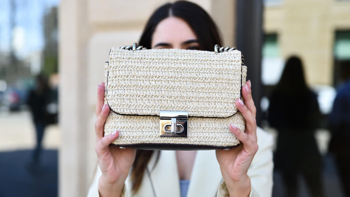 Person holding a textured handbag in front of their face with a blurred outdoor background